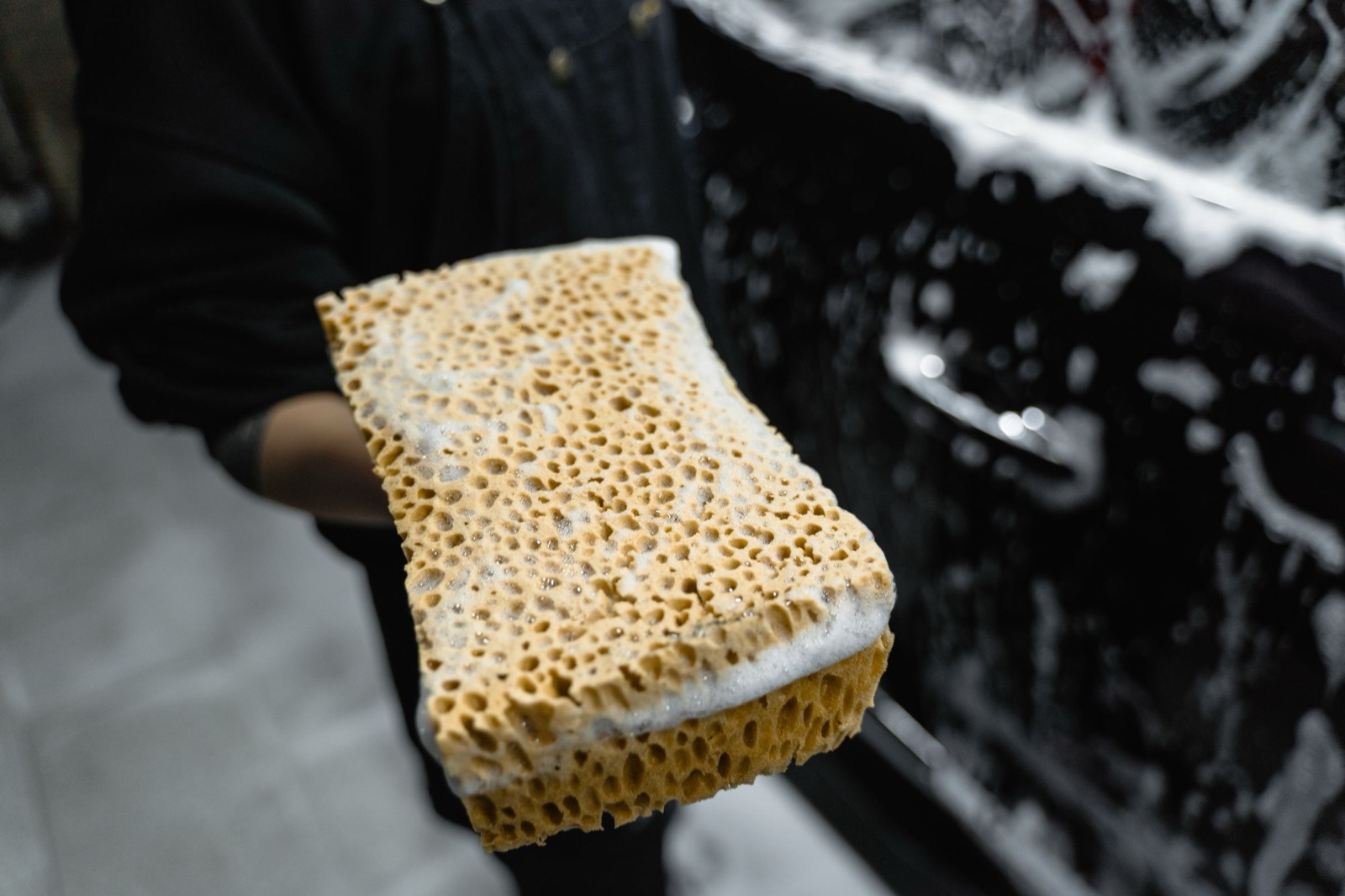 A foam-loaded wash sponge held up in front of a black car door covered in suds